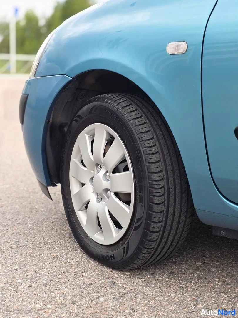 Sivulta: Vasemman etupuolen lähikuva. Sininen Renault Clio on pysäköity asfaltille. Kuvassa näkyy edessä oleva pyörä, jossa on hopeanvärinen teräkuppi ja musta renkaat.