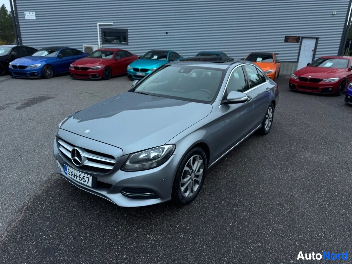 Etuviisto, nokka vasemmalle: Harmaa Mercedes-Benz C -sedan etukulmasta lähikuvana, keula vasemmalla, taustalla halli ja muita autoja; rekisterikilpi.