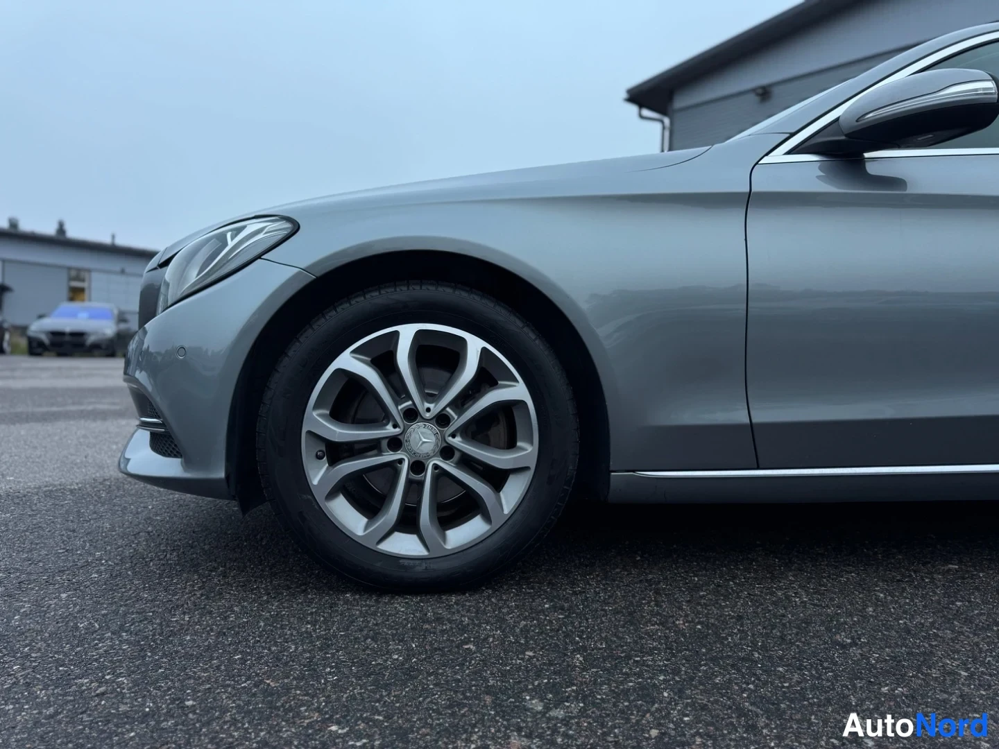 Etuviisto, nokka oikealle: Lähikuva auton vasemman etupään ja renkaasta, jossa on kromatut kehykset. Auto on harmaansävyinen, ja pinnan kiilto näkyy valossa.