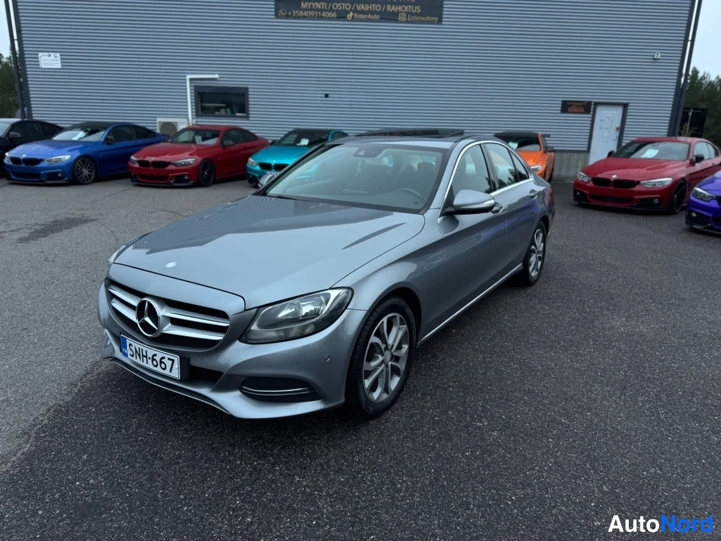 Etuviisto, nokka vasemmalle: Harmaa Mercedes-Benz C -sedan etukulmasta, keula vasemmalla, liiketilan pihalla; rekisterikilpi.