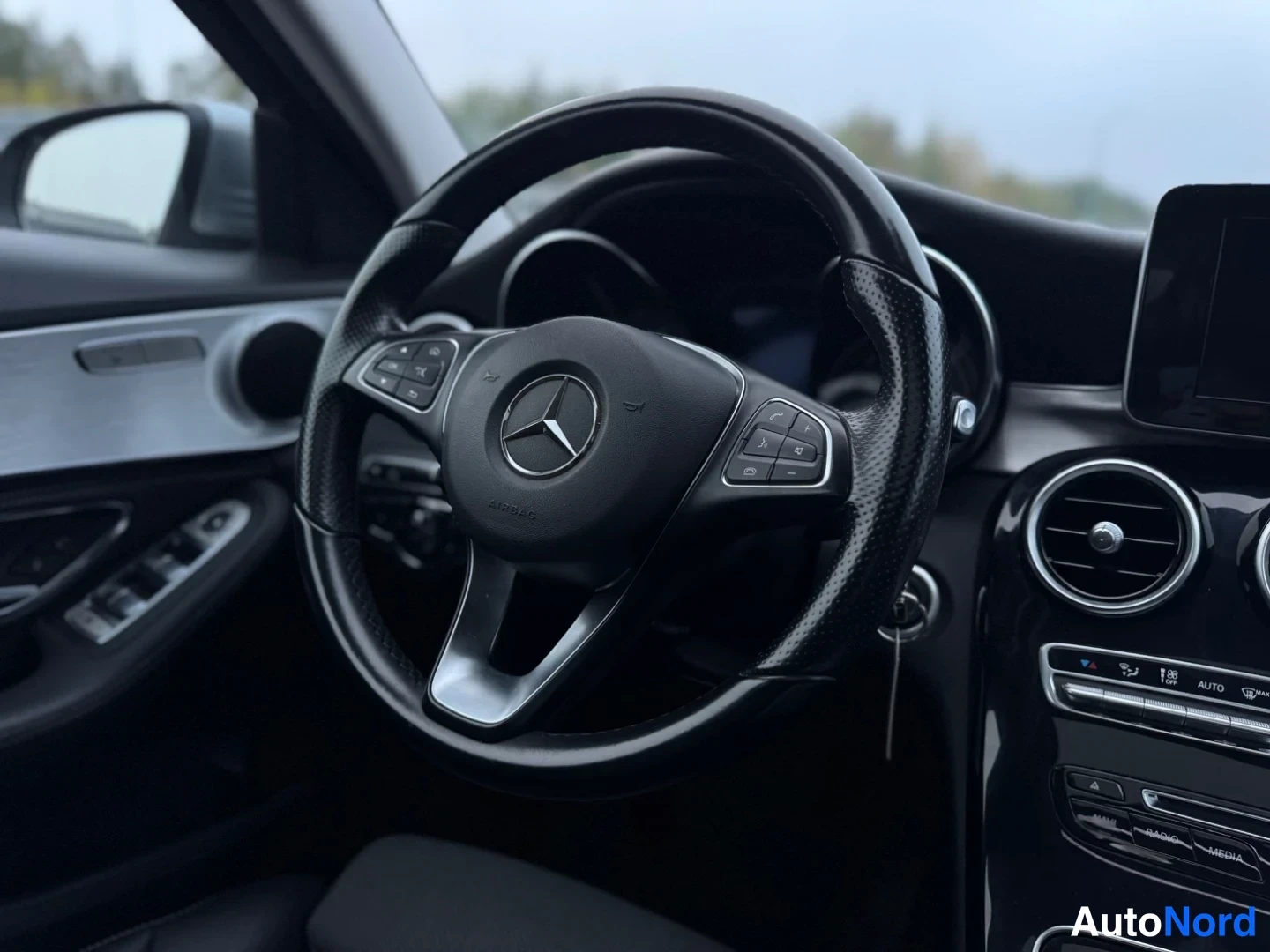 Edestä: Lähikuva Mercedes-Benz-ohjauspyörästä ja kojelaudasta; oven kytkimet ja osa keskikonsolin ilmastoinnista ja näytöstä näkyvät.