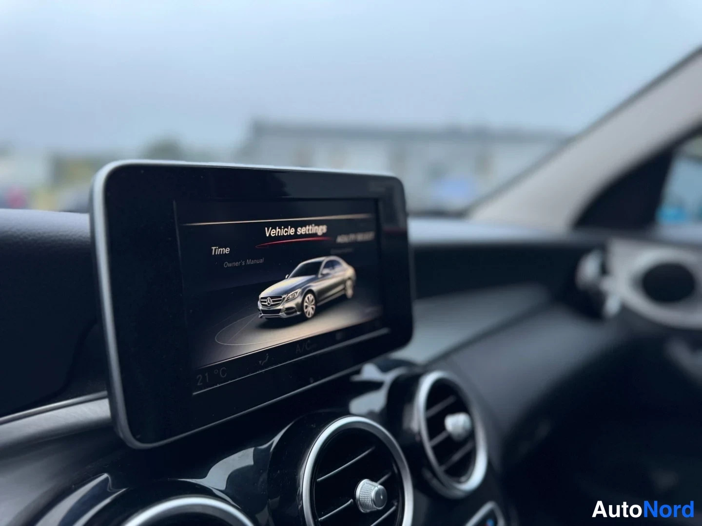 Edestä: Kojelaudan näyttö lähikuvaus: näytöllä teksti Vehicle settings, Time ja Owner's Manual sekä hopeisen auton kuva, alapuolella pyöreät ilmastointiaukot.