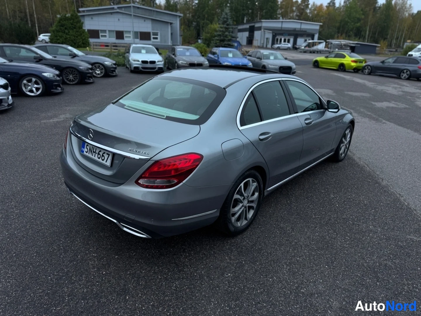 Takaviisto: Harmaan Mercedes-Benz C-sarjan sedan taka-vasen kolmasosa. Auto on parkkipaikalla; taustalla muita autoja ja rakennus.