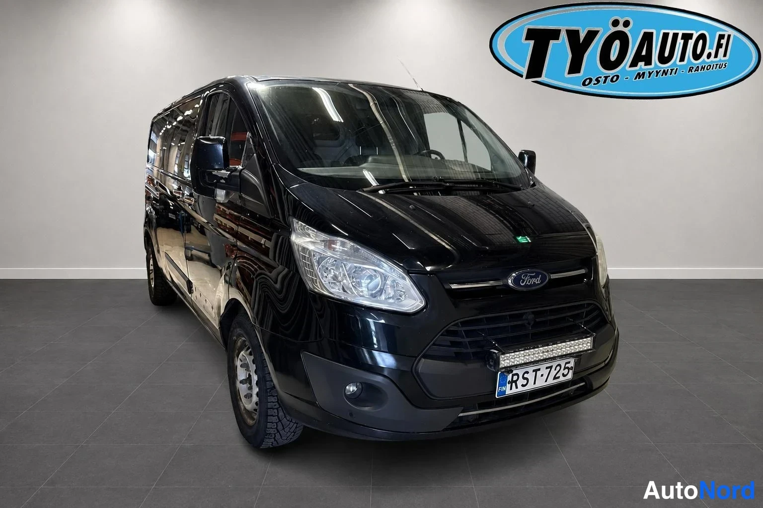 Etuviisto, nokka oikealle: Musta Ford Transit Custom -pakettiauto. Kuvassa auton etuosa ja vasen kylki näkyvät. Etupuskurissa on lisävalopalkki. Rekisterinumero