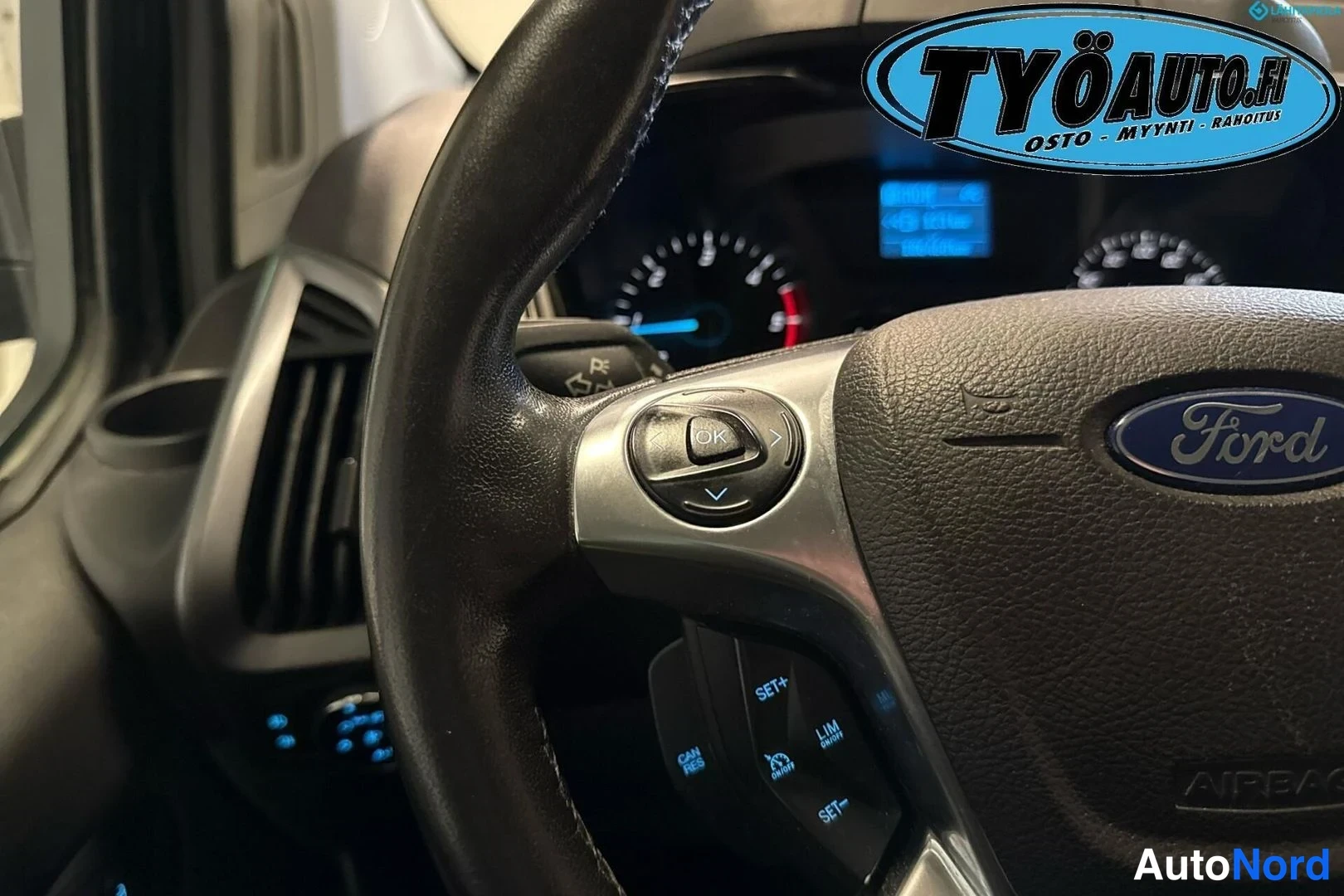 Ohjaamo: Steering wheel with Ford logo and multifunction buttons including cruise control (SET+, SET-, CANCEL) on the left spoke and trip computer navigation keys on the