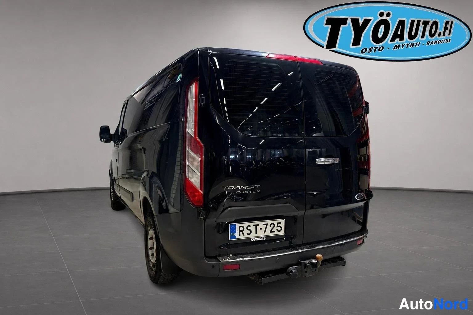 Takaviisto: Tummaansininen tai musta Ford Transit Custom pakettiauton takakulma. Takapuskuriin kiinnitetty vetopylväs (tow bar). Auto seisoo graniittilaatoilla sisätiloissa