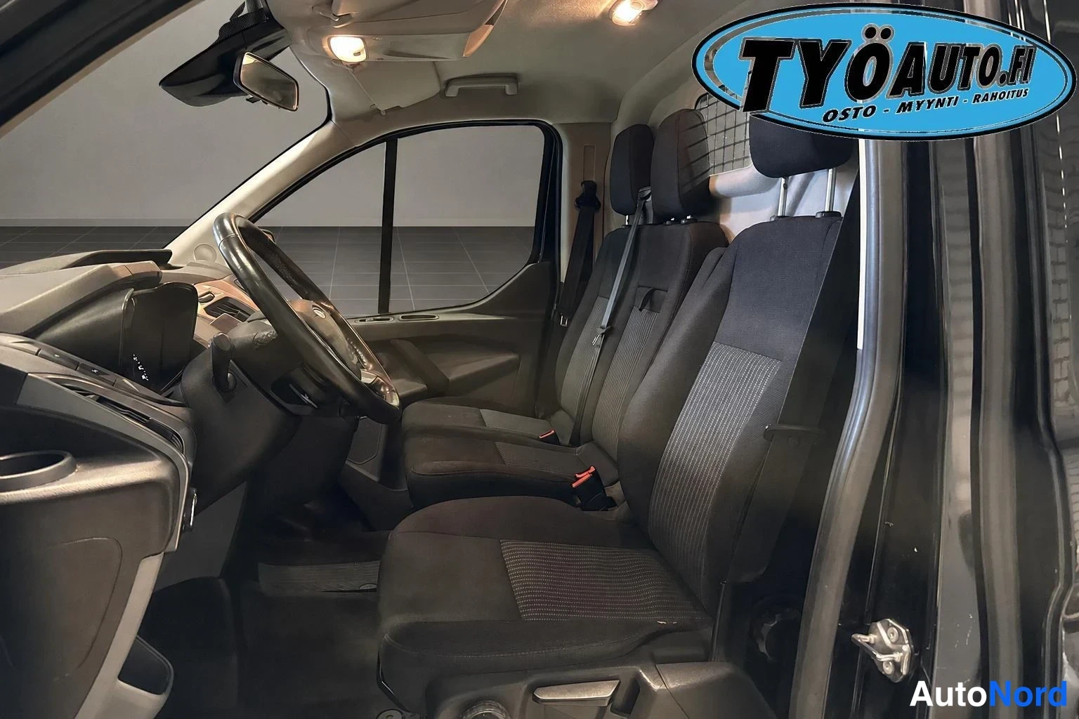 Ohjaamo: Ford Transit Customin edustan näkymä sivusta, kolme istuinta: kuljettajan ja matkustajan istuin sekä lisäistuin takaosan takana. Ohjauspyörä ja instrumenttirivi