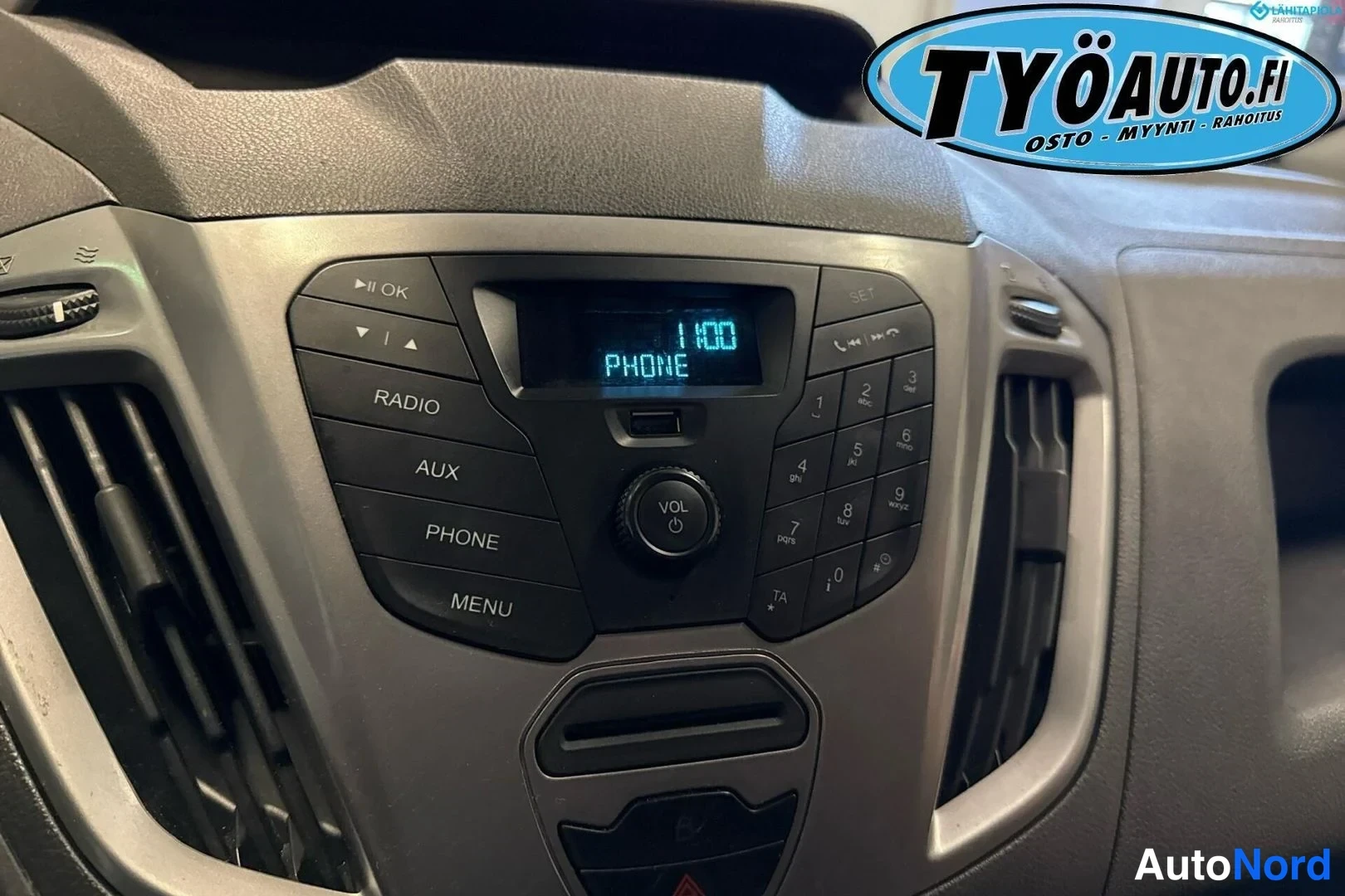 Ohjaamo: Dashboard close-up showing the central audio panel with a blue display reading '1:00 PHONE' surrounded by radio controls and a USB port.