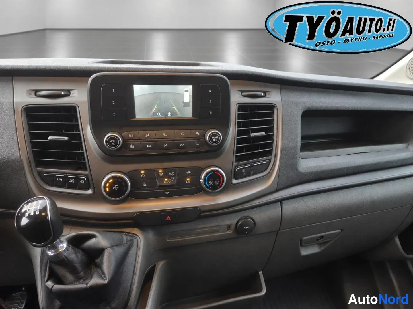 Yksityiskohta: Center console of the dashboard showing the infotainment screen, climate controls with rotary dials, and manual gear lever.