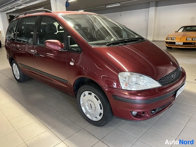 Etuviisto, nokka oikealle: Punaruskea tila-auto etukolmivartta oikealta, kattotelineet ja hopeiset pyöränkannattimet näkyvät, autosalon lattialla.