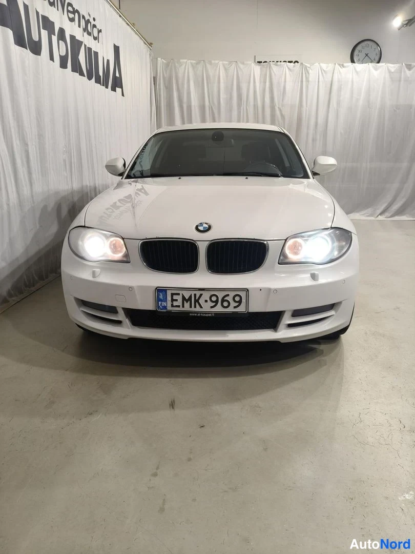 Edestä: Valkoisen BMW:n etunäkymä suoraan edestä, ajovalot päällä, rekisterikilpi.