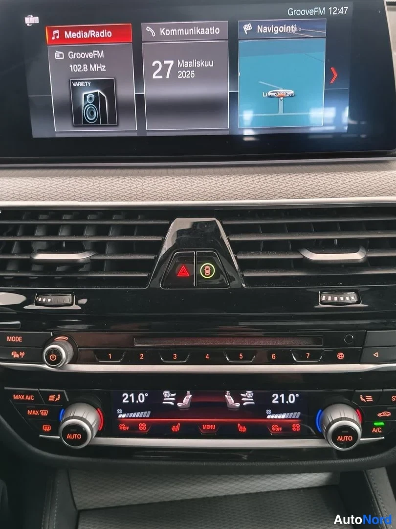Muu: Keskikonsolin näyttö ja automaattinen ilmastointipaneeli; näytössä 'GrooveFM 102.8 MHz', '27 Maaliskuu 2026' ja 'Navigointi'; molemmat 21.0 astetta AUTO.