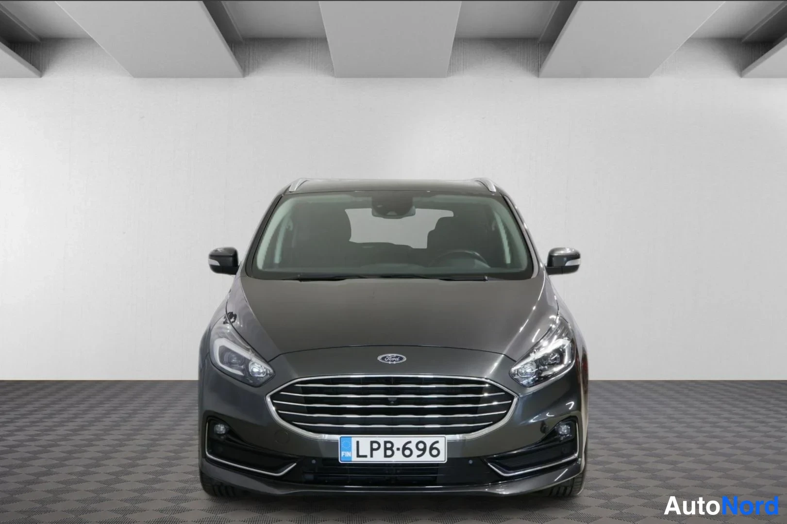 Edestä: Edessä kuvattuna hopeanharmaa Ford S-Max. Etuosassa on Ford-logon ja grillin, sekä LED-päävalojen sijaintipaikka. Takana on rekisterinumero.