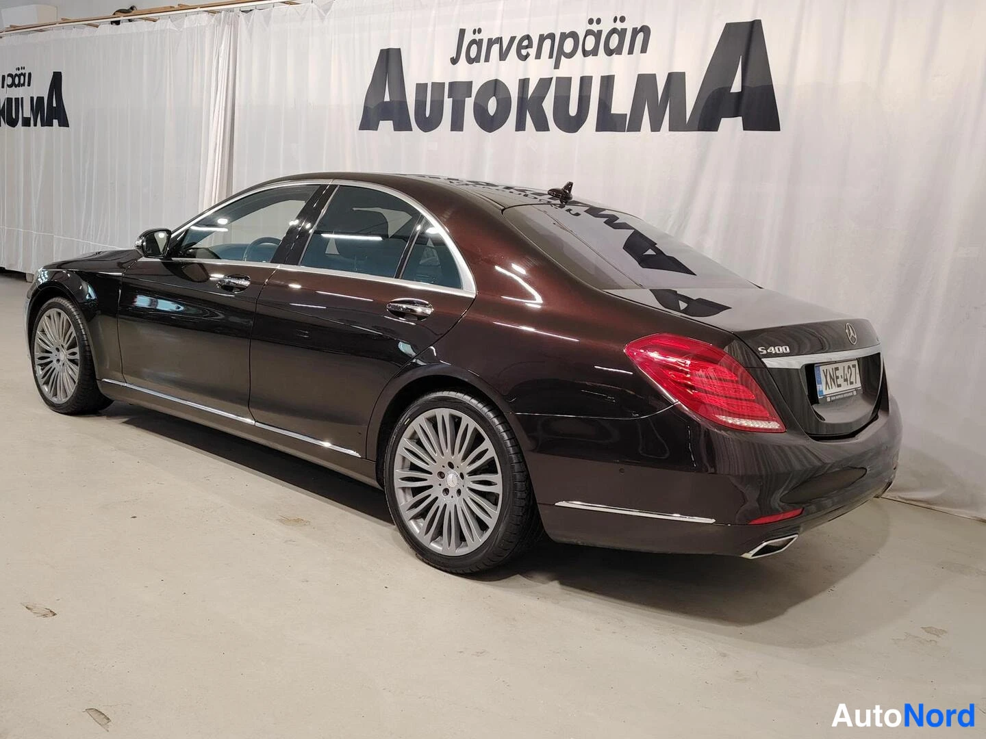 Takaviisto: Tummanruskean Mercedes-Benz S 400 -sedanin takakulmanäkymä oikealta, rekisterikilpi, kuvattuna sisätiloissa valkoista Autokulma-taustaverhoa vasten.