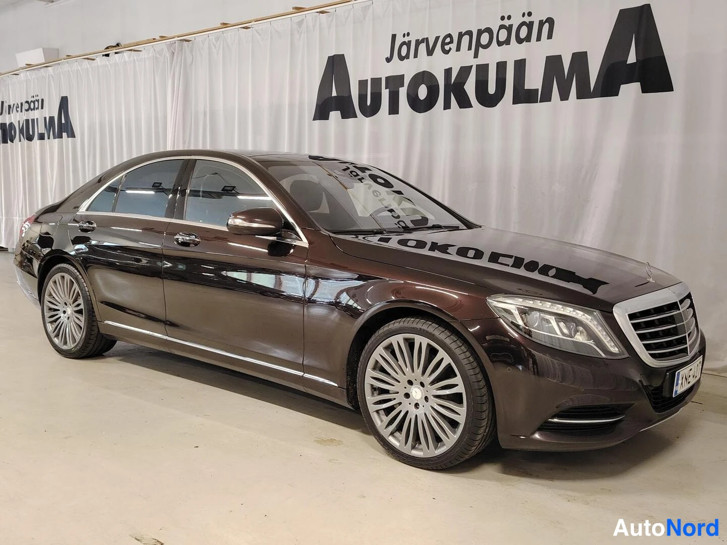 Etuviisto: Tummanruskean Mercedes-Benz S 400 -sedanin etukulmanäkymä oikealta, kroomiset etusäleikön kehykset ja kevytmetallivanteet näkyvissä, kuvattuna sisätiloissa.