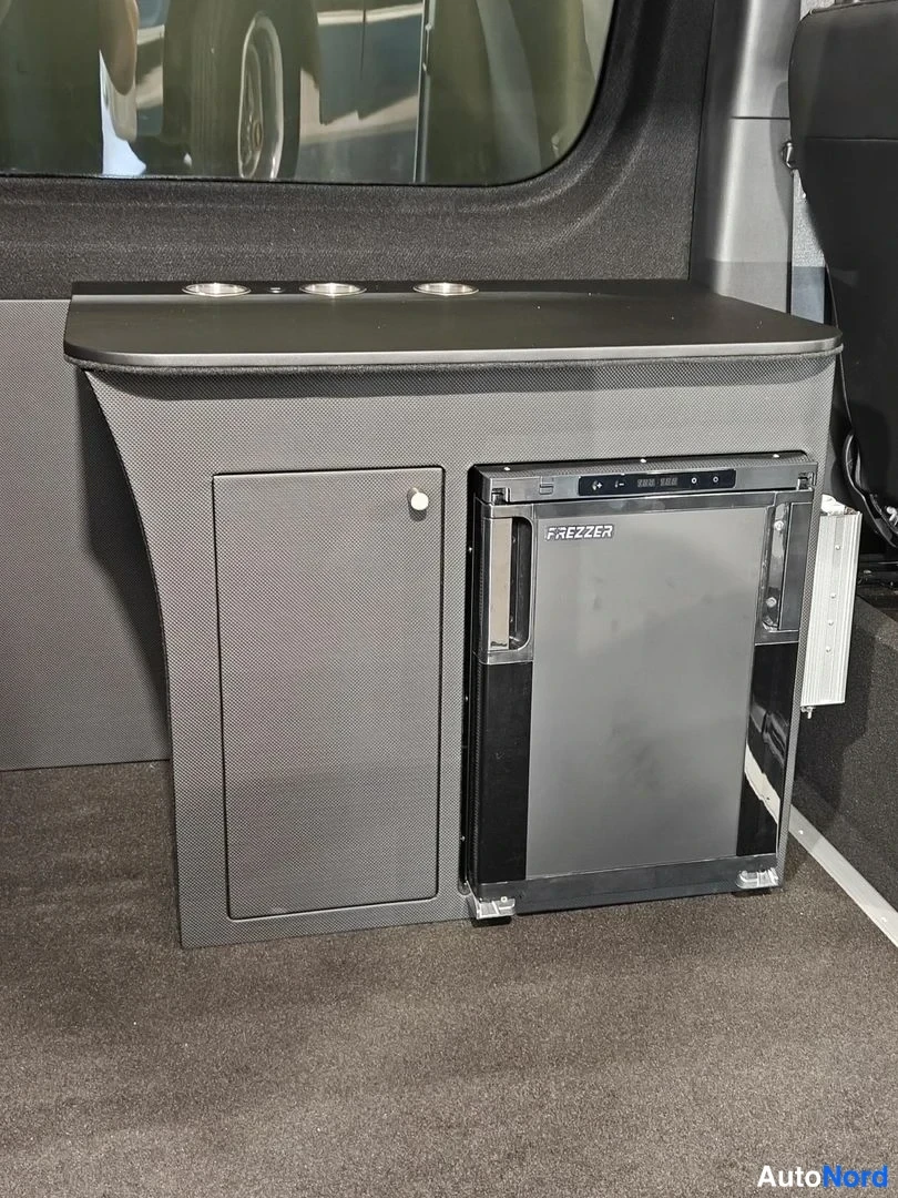 Muu: Auton sisätilassa tumma kaapimainen kommodi, jossa on jalustalle asennettu musta FREEZER-jäähdytyslaatikko ja työtaso, jossa on kolme metallista kuplipidikettä.