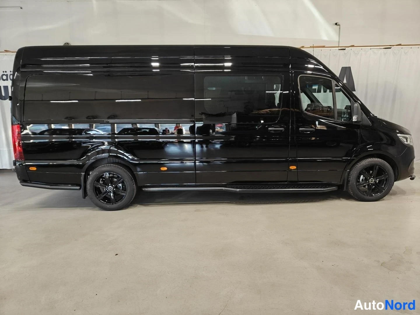 Sivulta: Musta Mercedes-Benz Sprinter -pakettiauto valkoisessa tilassa profiilikuvassa. Takajalkakoskettimet ja mustat kevytmetallivanteet näkyvät.