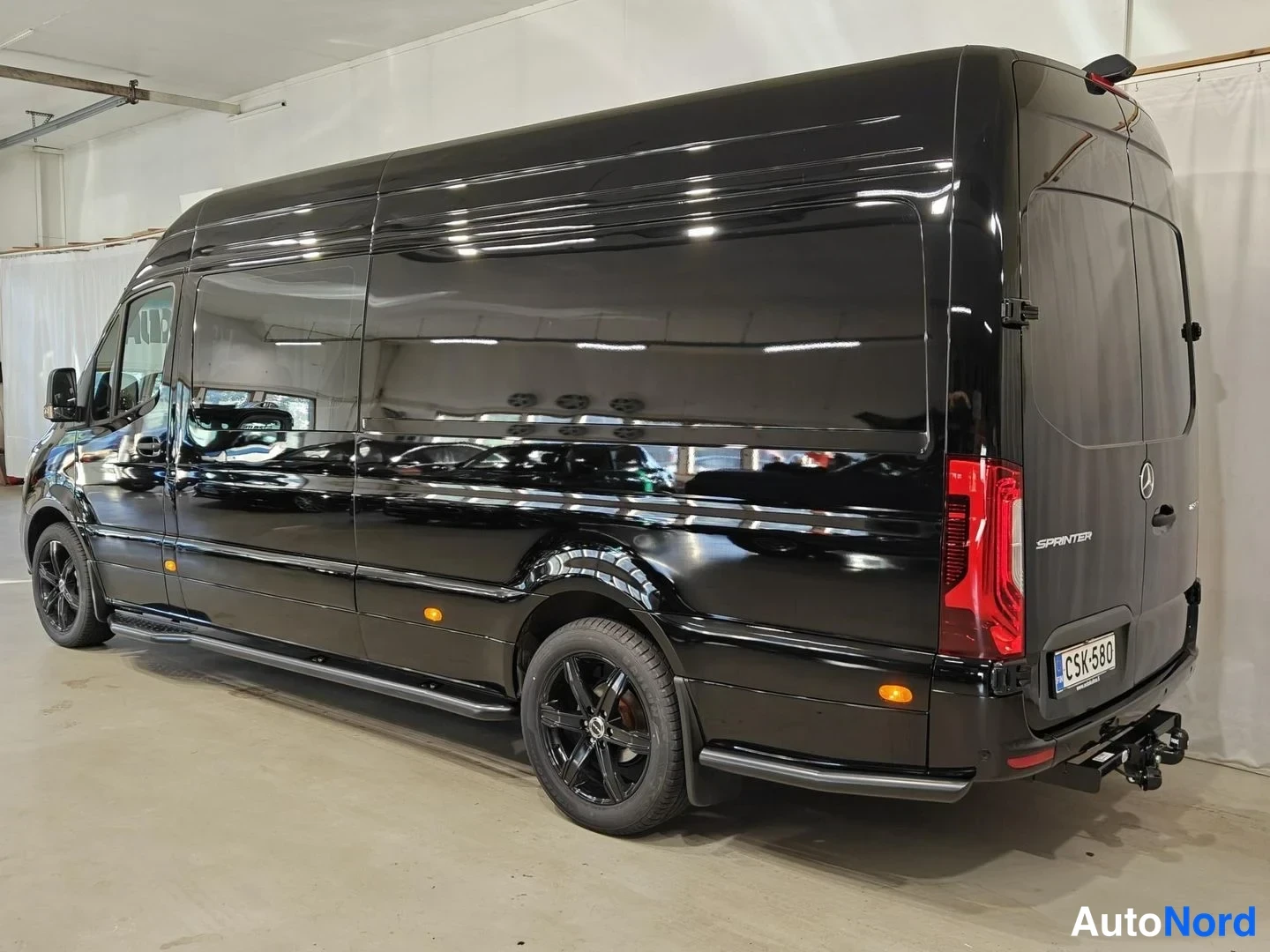 Takaviisto: Musta Mercedes-Benz Sprinter -pakettiauto takana kolmen neljänneksen kulmasta. Rekisterinumero ja perämittari selkeästi näkyvissä.