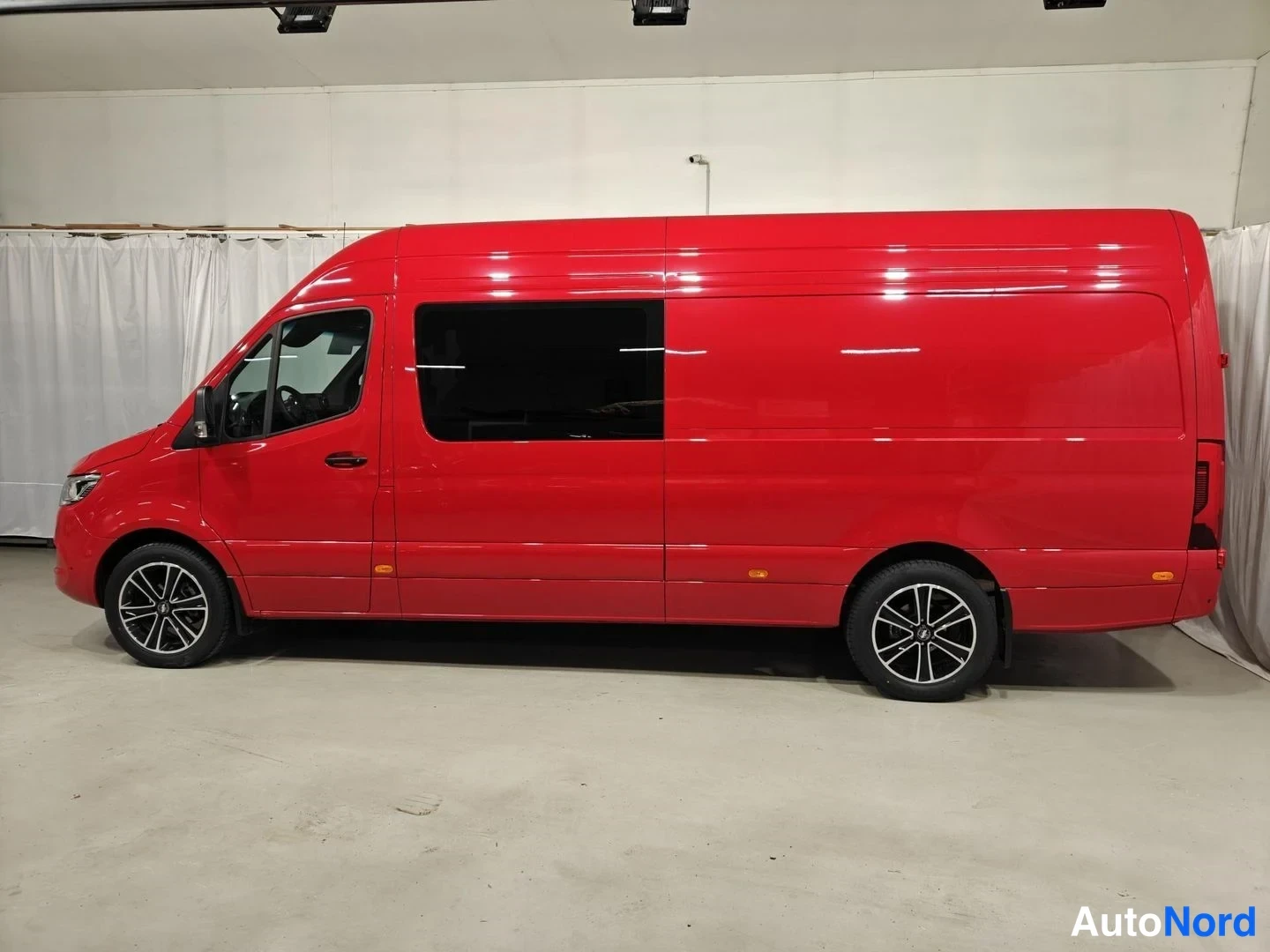 Sivulta: Punainen Mercedes-Benz Sprinter -pakettiauto sivusta sisätiloissa, pitkä korirakenne ja tummat ikkunat.