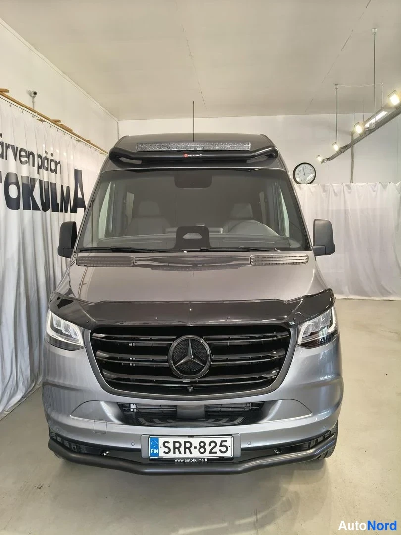 Edestä: Suoraan edestä kuvattu harmaa Mercedes-Benz -pakettiauto, rekisterikilpi, musta griffikehys ja katonäytön kaltainen lisävalo.