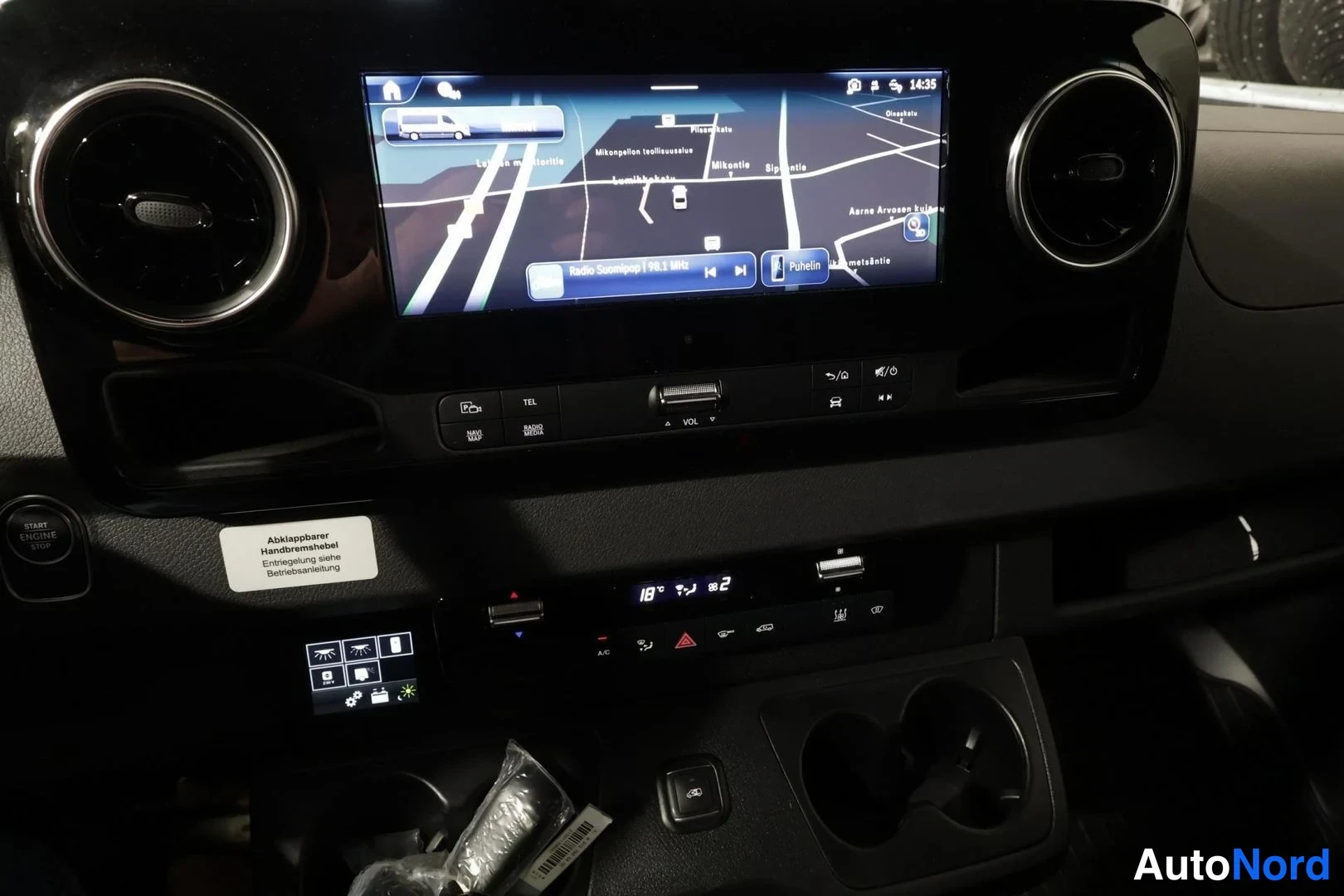 Yksityiskohta: Mercedes-Benzin kojelaudan keskikonsoli: kosketusnäyttö, joka näyttää navigointikartan ja radiosoittimen, ympärillä pyöreät ilmastointisuuttimet, alla ilmastoin