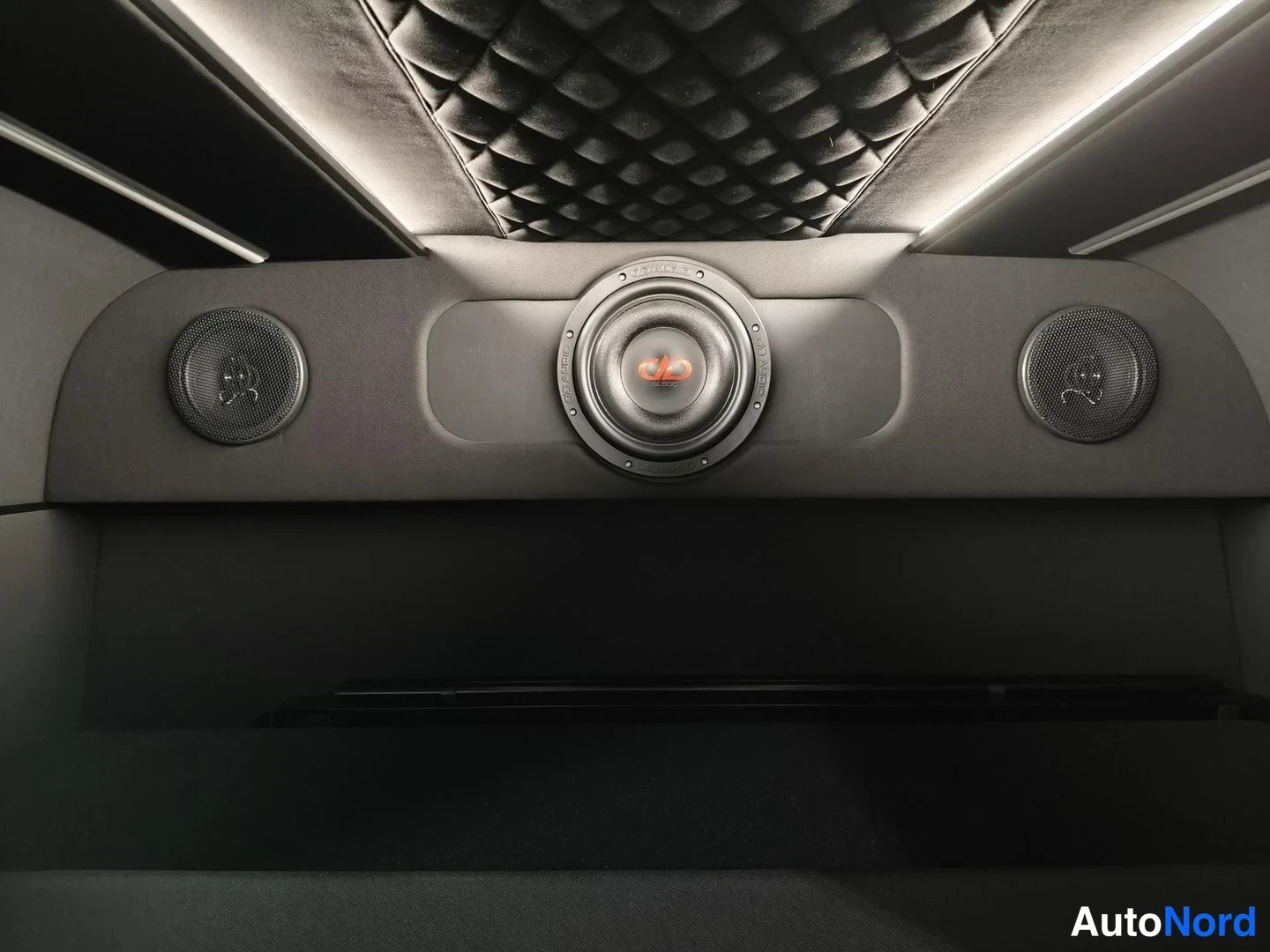 Muu: Auton sisätiloissa nähnyt paneeli, jossa on iso DD Audio -subwoofer ja kaksi pientä kaiutinta molemmin puolin, tummaa verhoiltu katto ja kotelot yläpuolella.