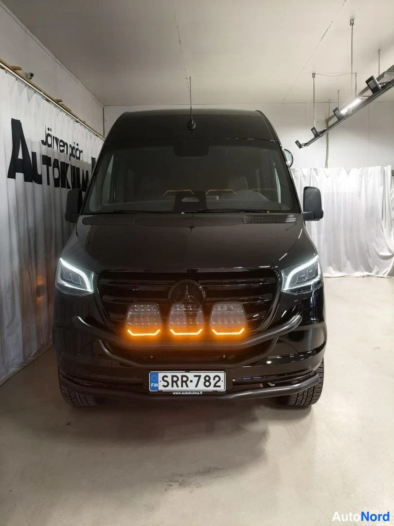 Edestä: Mustan pakettiauton etunäkymä sisätiloissa; etusäleikössä kolme oranssivaloista lisävaloa, rekisterikilpi.