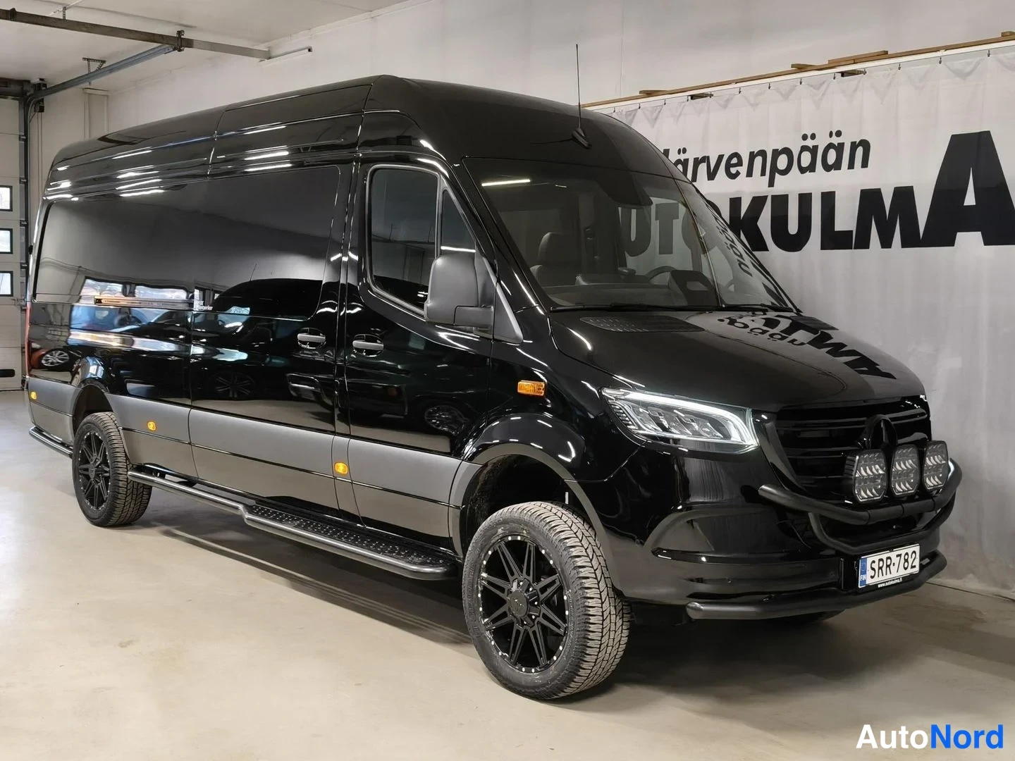 Etuviisto, nokka oikealle: Musta Mercedes-Benz Sprinter -pakettiauto etuviistosta kuvattuna sisätilassa.