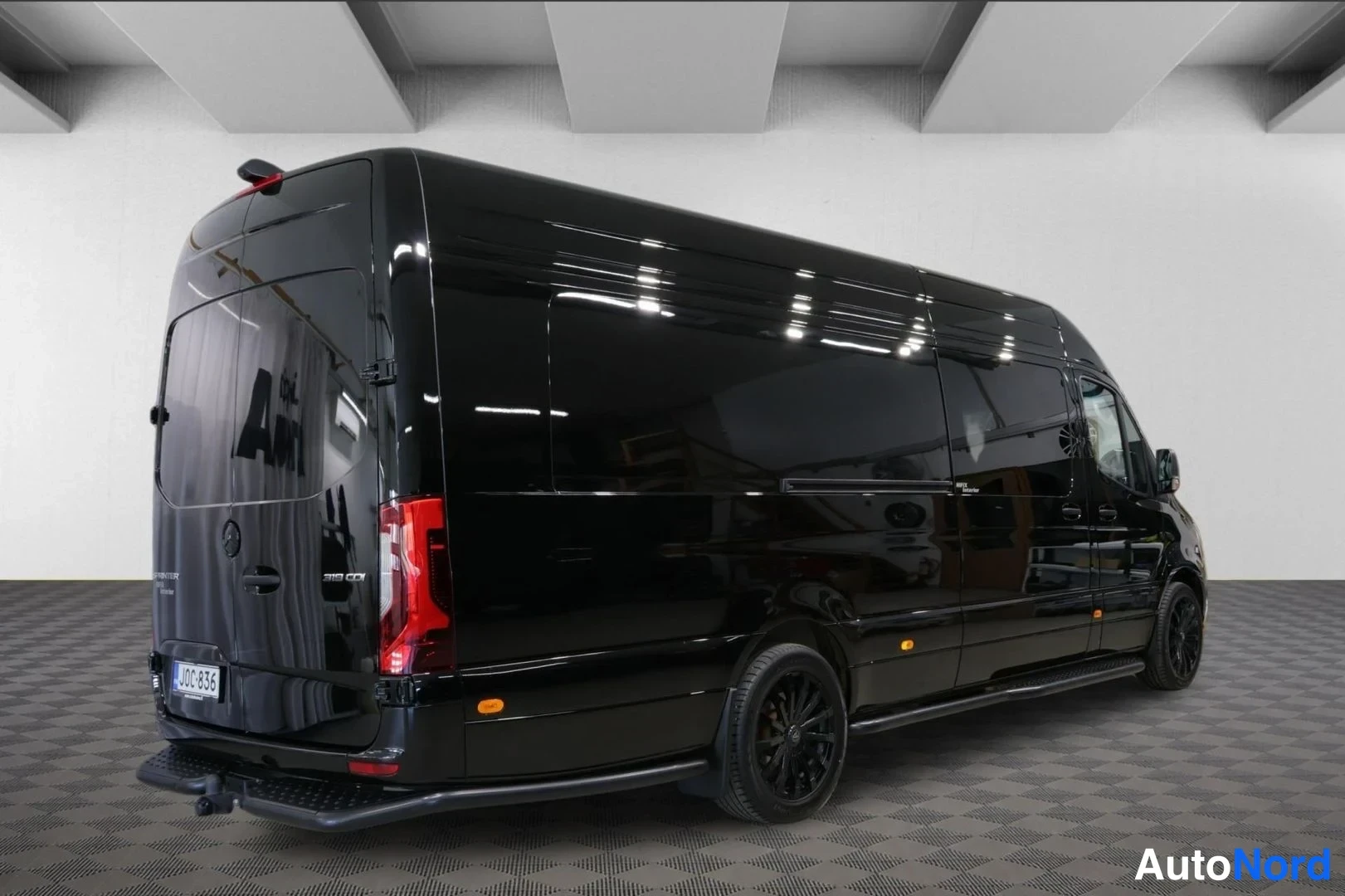 Takaviisto: Musta Mercedes-Benz Sprinter -pakettiauto takaa viistosta, korkea pitkä korimalli, tummat vanteet ja vetokoukku näkyvissä.