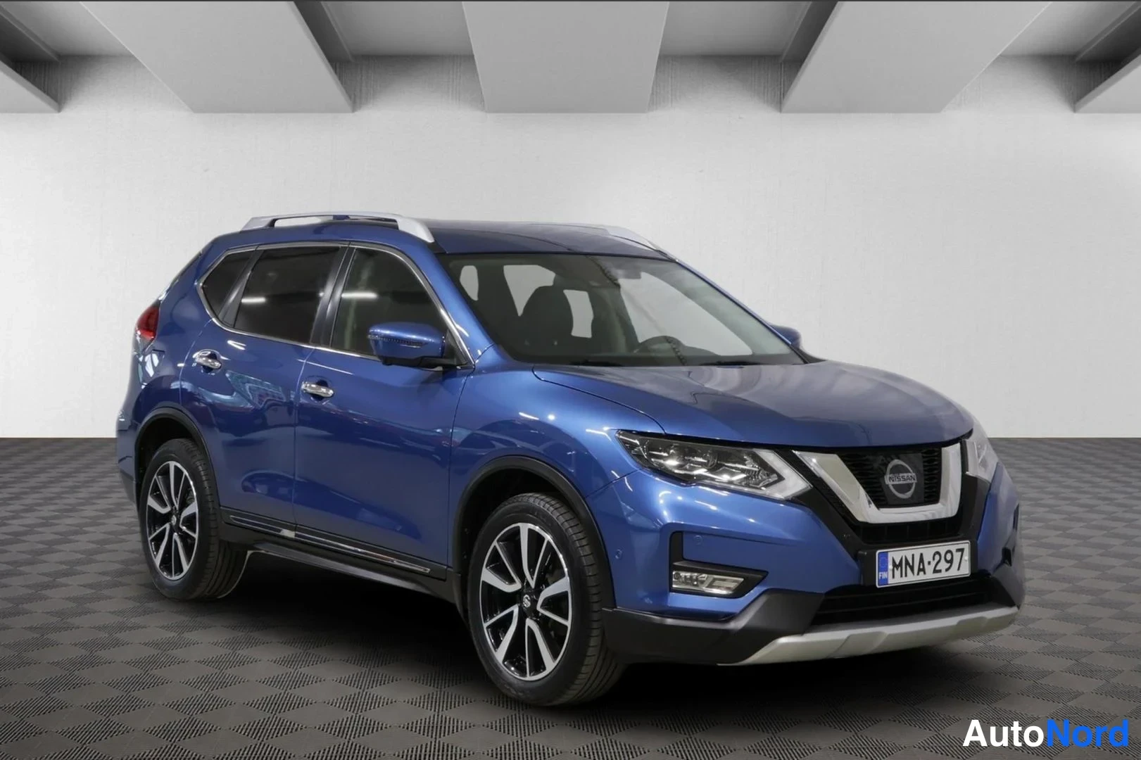Etuviisto, nokka oikealle: Sininen Nissan X-Trail -maasturi etukulmasta oikealta kuvattuna sisätiloissa, rekisterikilpi näkyvissä.