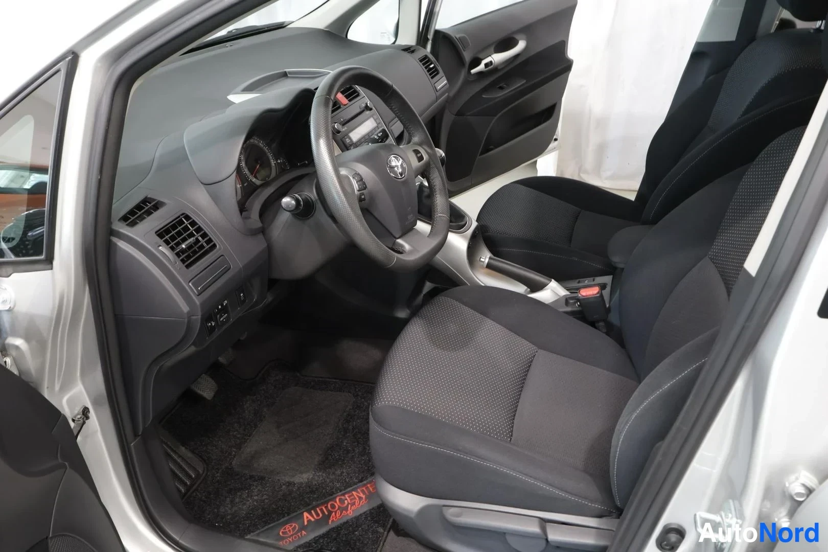 Muu: Auton sisätilat ohjaamosta takaosan suuntaan: Toyota-ohjauspyörä, kojelauta, kangasistuimet ja AUTOCEnter-merkkiset lattiamatot.