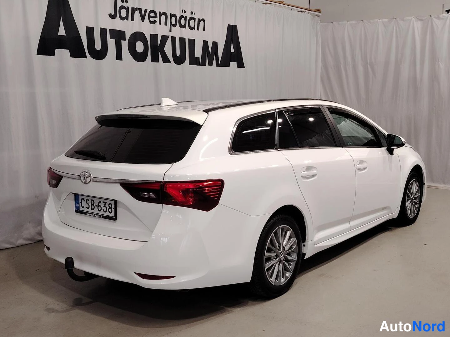 Takaviisto: Valkoisen Toyota Avensis -farmariauton takakulmanäkymä sisätiloissa, rekisterikilpi, taustalla Järvenpään Autokulma -tausta.