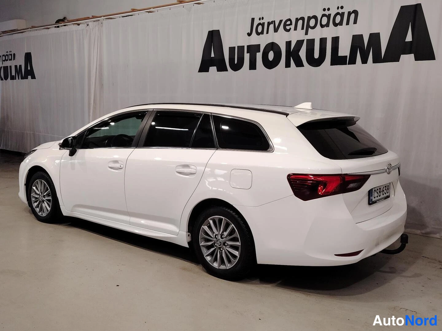 Takaviisto: Valkoisen Toyota Avensis -farmariauton takaosa ja vasen kylki sisätiloissa, rekisterikilpi, taustalla Järvenpään Autokulma -tausta.