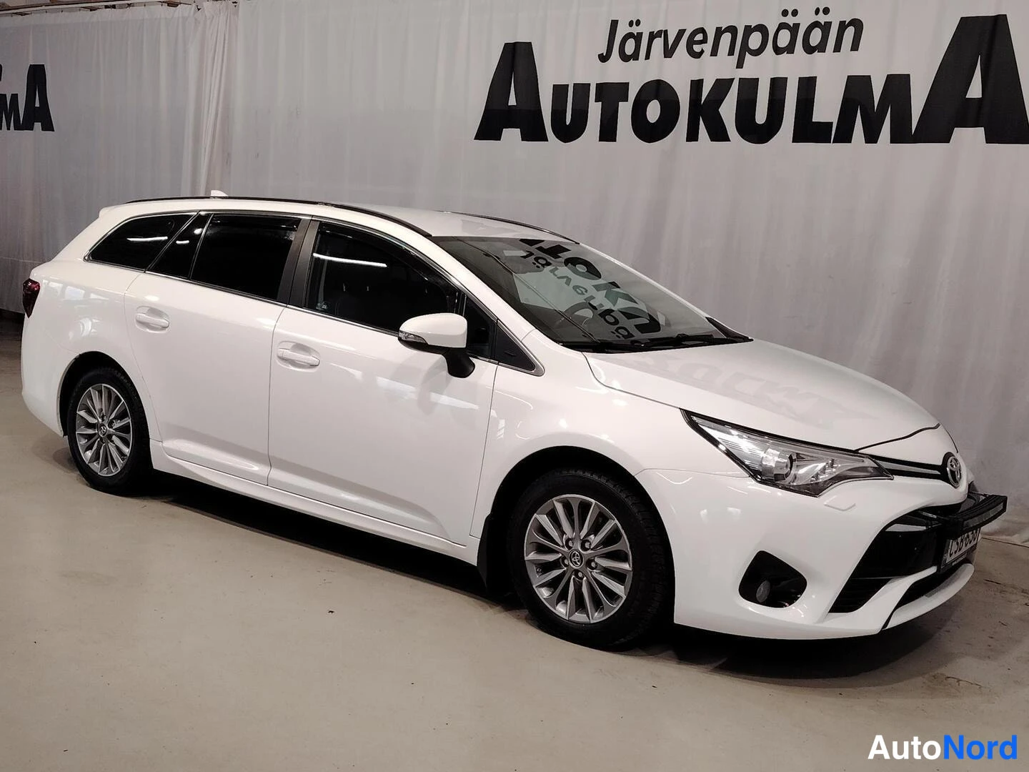 Etuviisto, nokka oikealle: Valkoisen Toyota Avensis -farmariauton keulakulmanäkymä sisätiloissa, keula oikealla, vasen kylki ja kattotelineet näkyvät, taustalla Järvenpään Autokulm