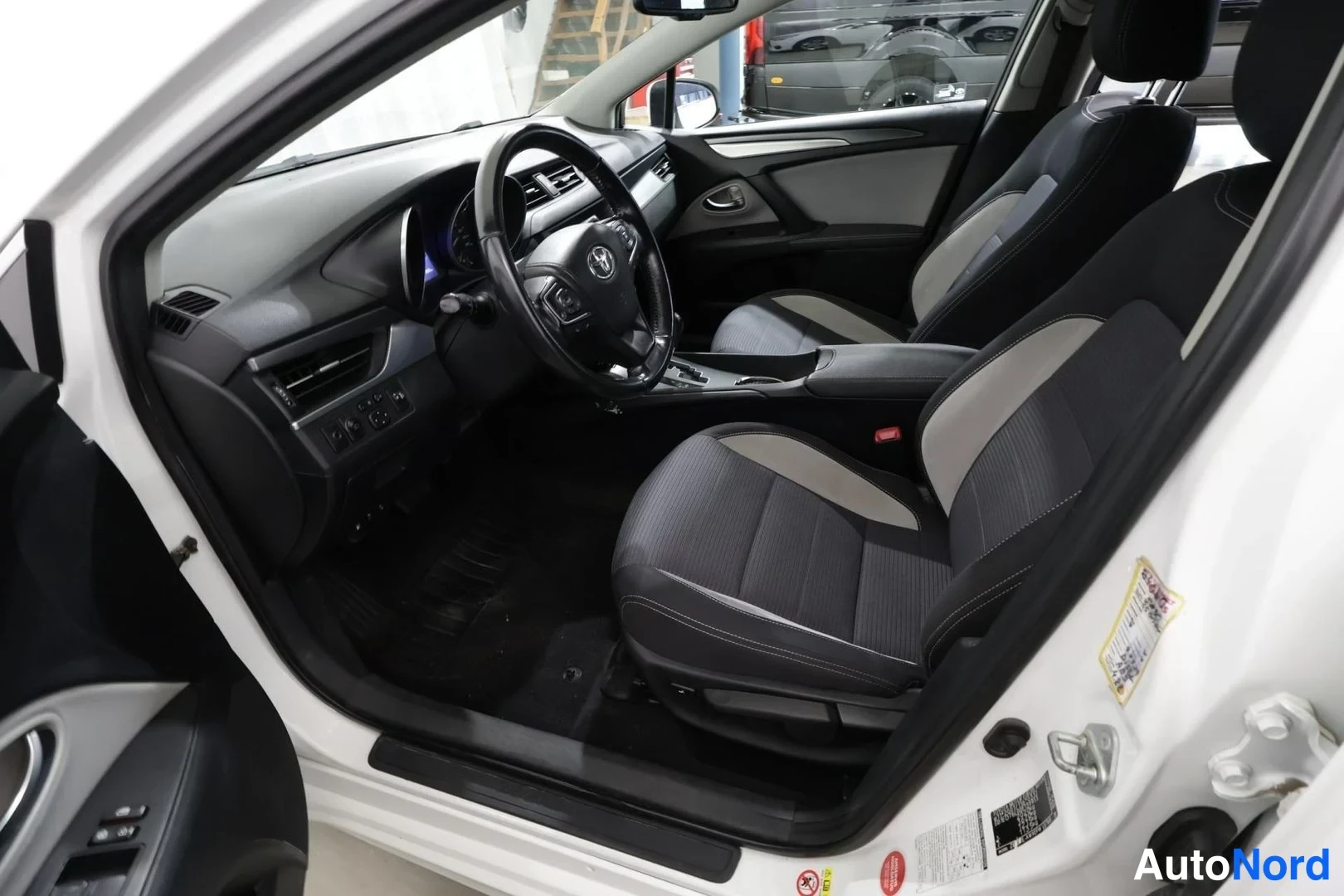 Sivulta: Avonaisen auton kuljettajan puolen sisätilat: Toyota-ohjauspyörä, kojelauta, vaihteenvalitsin ja musta-harmaat penkit.