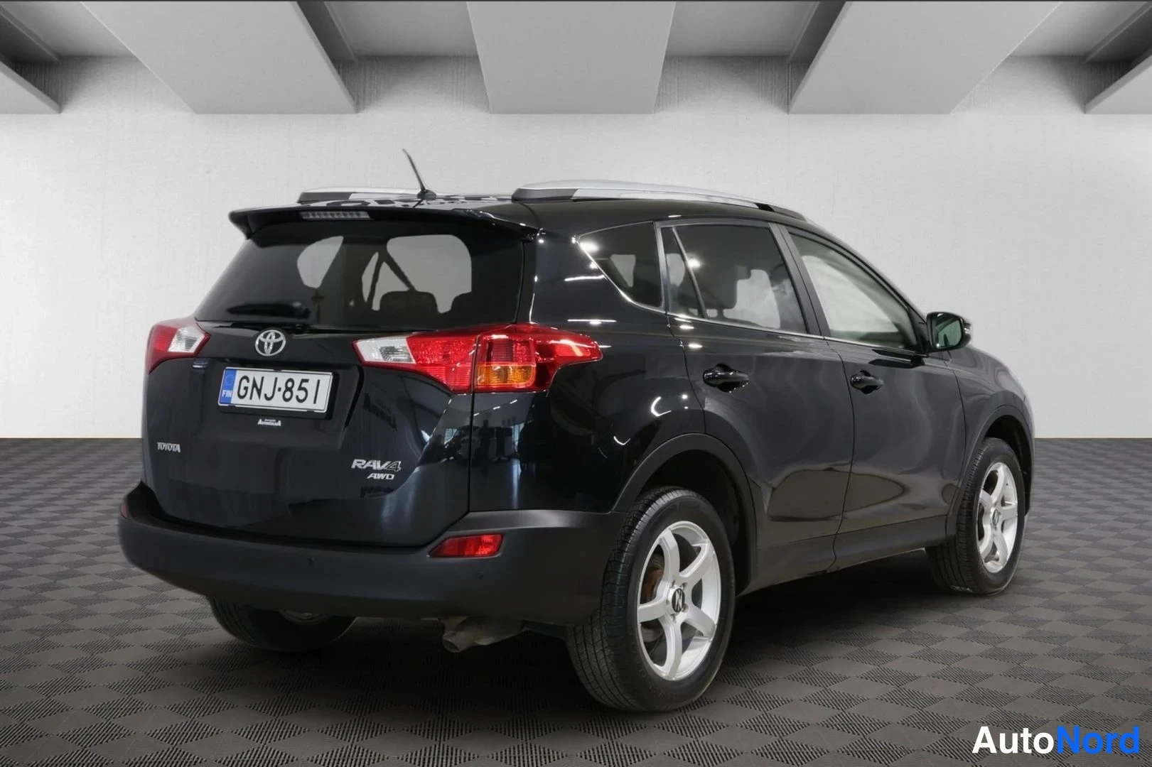 Takaviisto: Musta Toyota RAV4 takaviistosta oikealta; rekisterikilpi, kattotelineet ja hopeavanteet näkyvät.