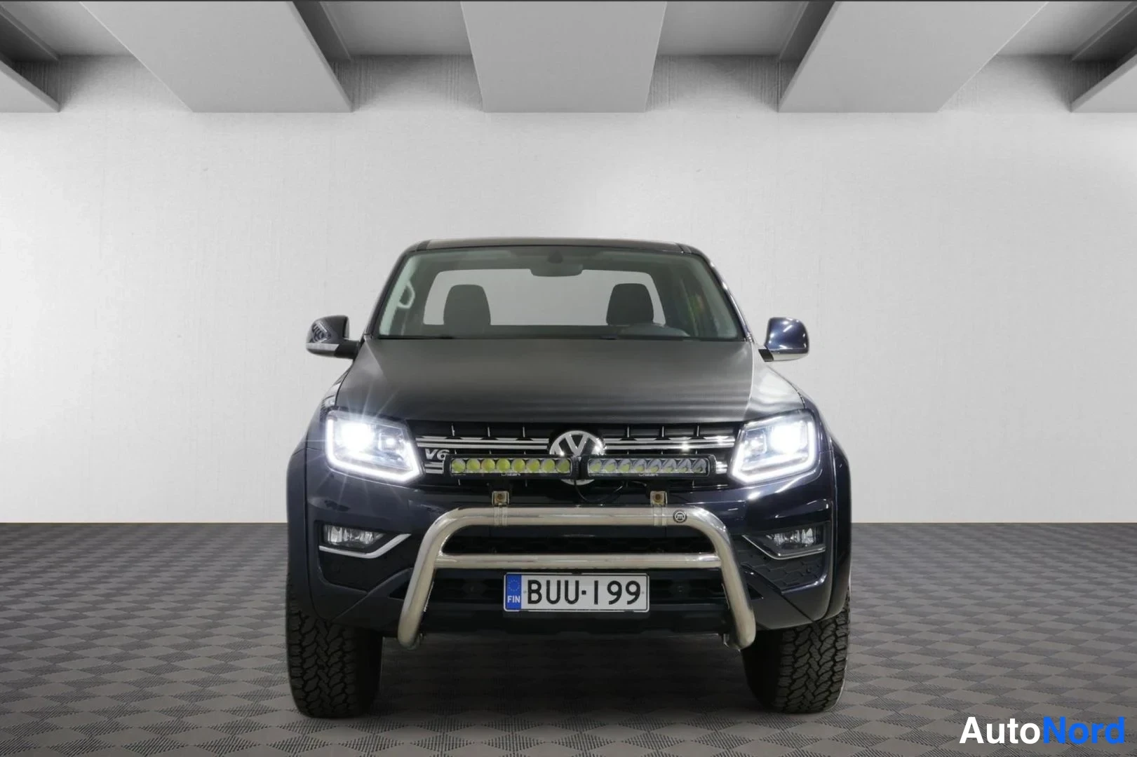 Edestä: Tummansinisen Volkswagen Amarok -lavauton etunäkymä; ajovalot päällä, kromikaari etupuskurissa ja lisävalokisko, rekisterikilpi.