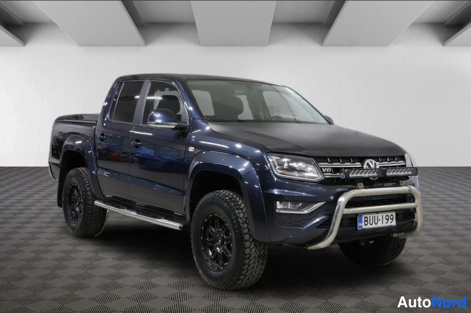 Etuviisto, nokka oikealle: Tummansininen Volkswagen Amarok -lava-auto vasemmalta etuviistosta; mustat kevytmetallivanteet, sivukaiteet ja kromikaari etupuskurissa.