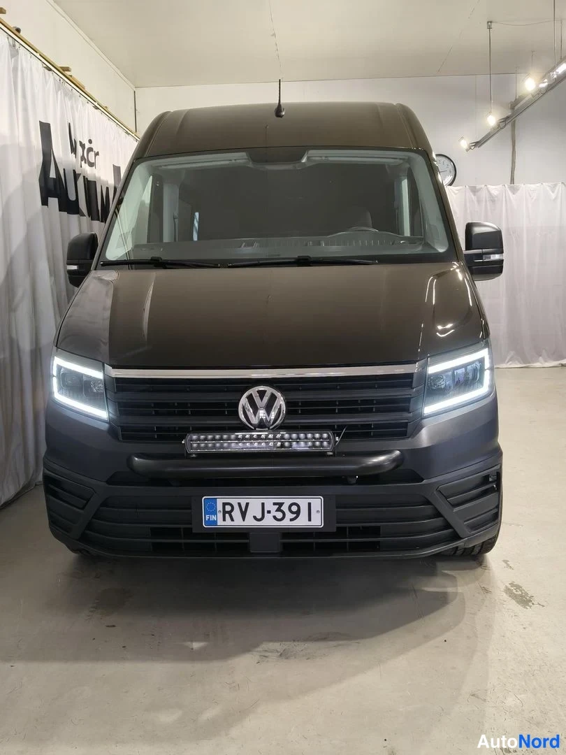 Edestä: Pakettiauton etunäkymä sisätiloissa, VW-merkki etusäleikössä, etivalot päällä, lisävalokisko puskurissa ja rekisterikilpi.