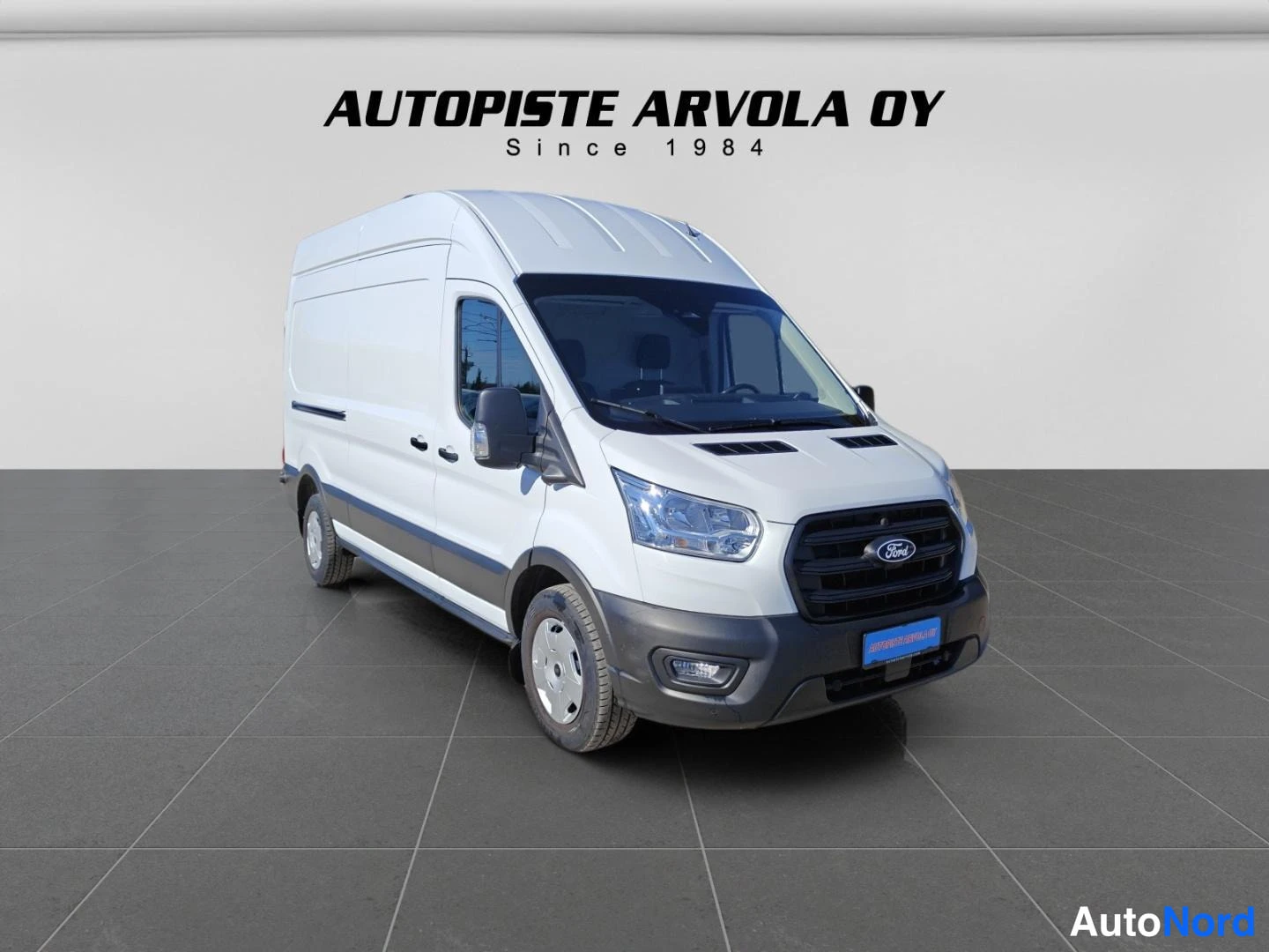 Etuviisto, nokka oikealle: Ford Transit 2025 - pakettiauto etuviistosta oikealle nojaavassa kulmassa, valkoinen kori, musta etusäleikkö ja puskurit, harmaa laatoitettu lattia ja va