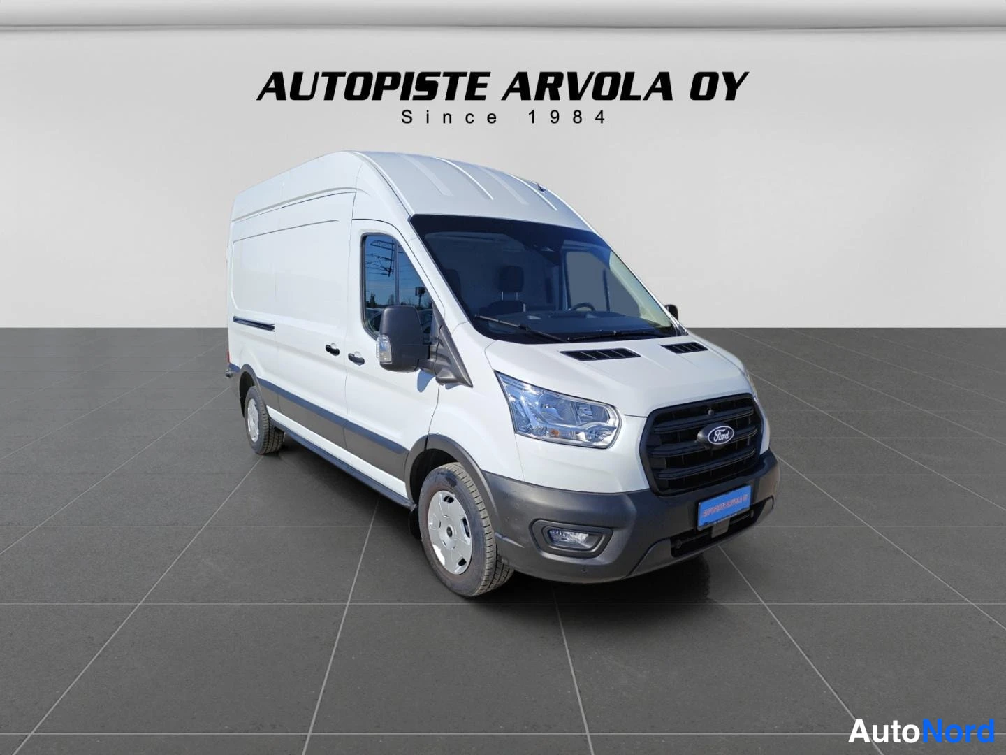 Etuviisto, nokka oikealle: Valkoinen Ford Transit -pakettiauto kolmasosakulmassa, keula ja vasen sivu näkyvissä; harmaa laatoitettu lattia ja vaalea tausta, taustalla teksti 'AUTOP