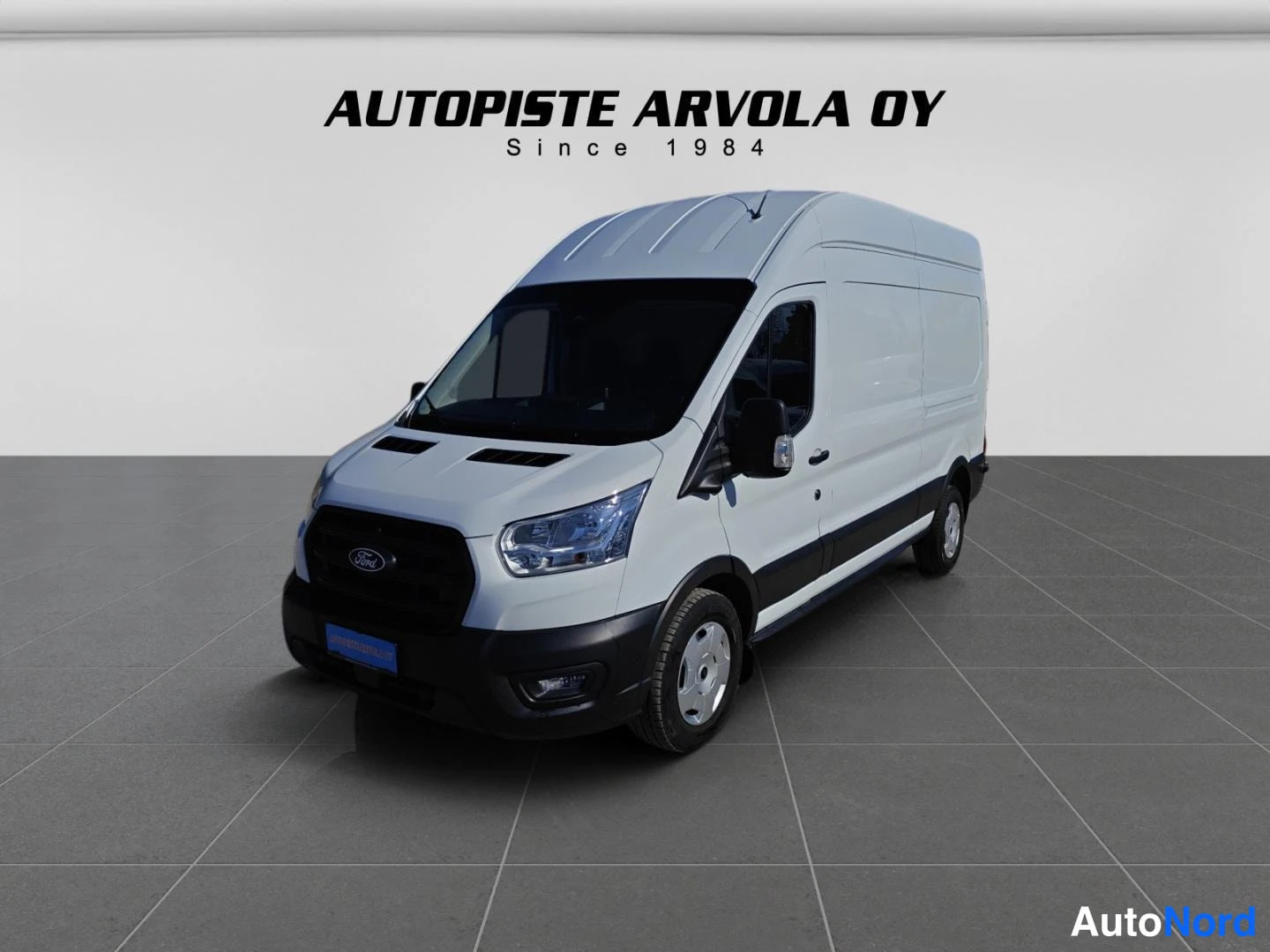 Etuviisto, nokka vasemmalle: Valkoinen Ford Transit -pakettiauto kolmasosakulmassa, keula ja oikea sivu näkyvissä; harmaa lattia ja vaalea tausta, taustalla teksti 'AUTOPISTE ARVOL