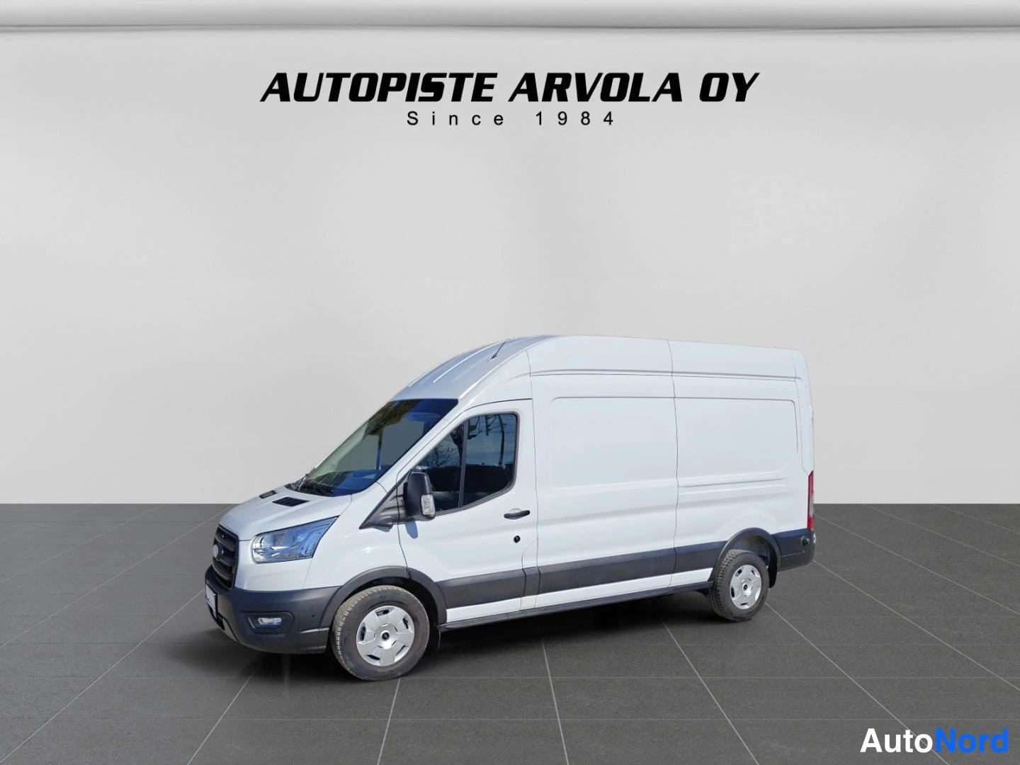 Etuviisto, nokka vasemmalle: Ford Transit 2025 - pakettiauto etuviistosta vasemmalle nojaavassa kulmassa korkealla katolla, valkoinen kori, tummat alapaneelit ja puskurit, harmaa l