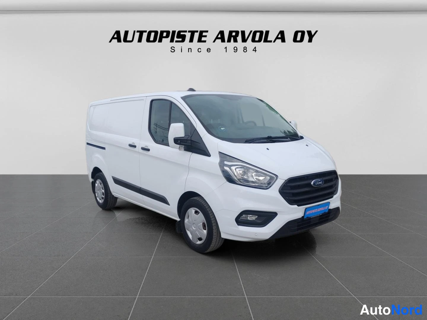 Etuviisto, nokka oikealle: Valkoinen Ford Transit Custom -pakettiauto kolmesta neljänneksestä edestä oikealta kuvattuna studiohallissa