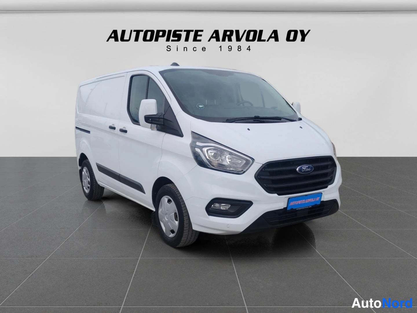 Etuviisto, nokka oikealle: Valkoinen Ford Transit Custom -pakettiauto kolmesta neljänneksestä edestä oikealta kuvattuna studiohallissa