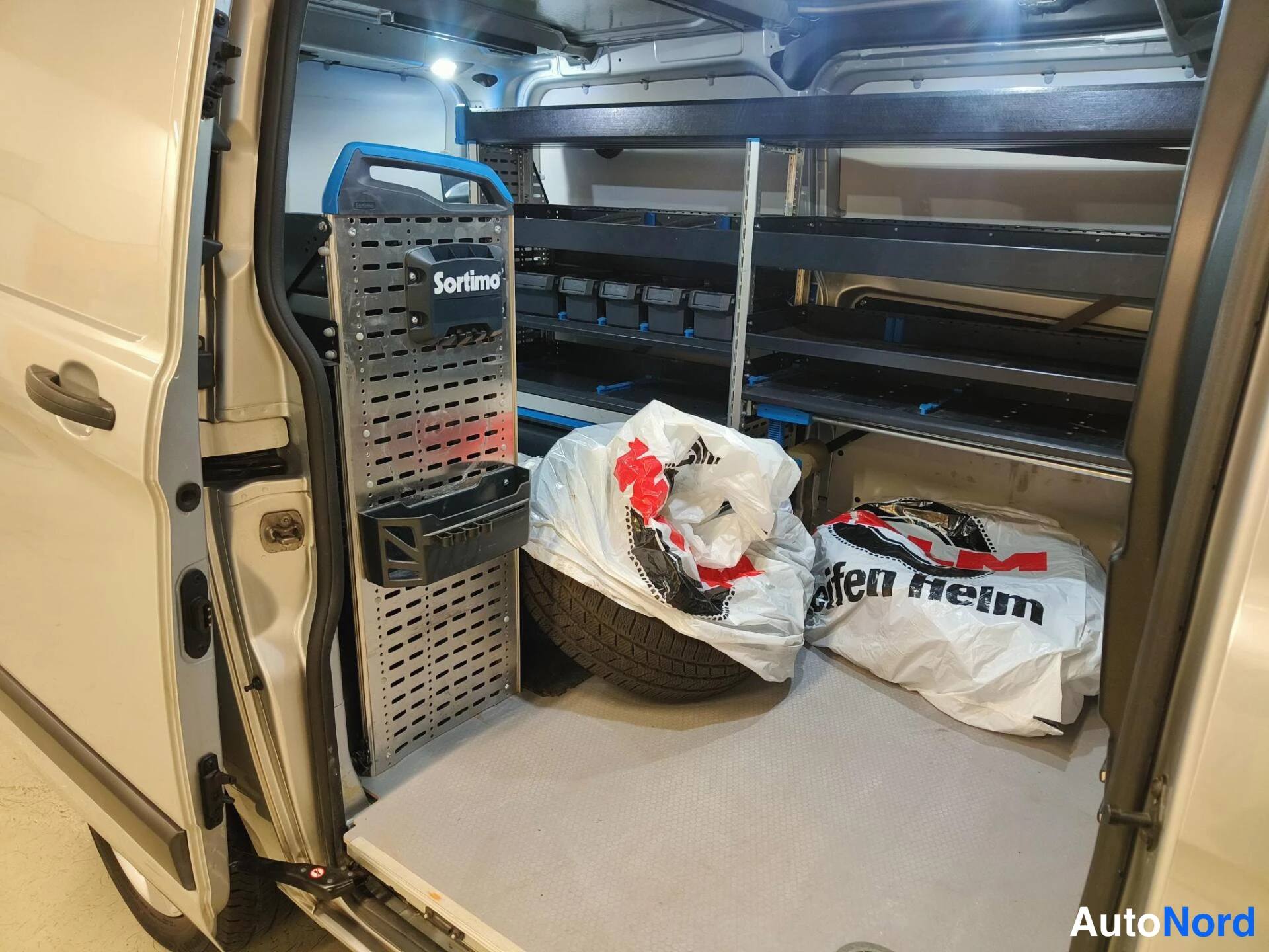 Muu: Pakettiautoon avoin takaluukku paljastaa kuormatilan Sortimo-hyllyineen; tilassa renas ja kassit.