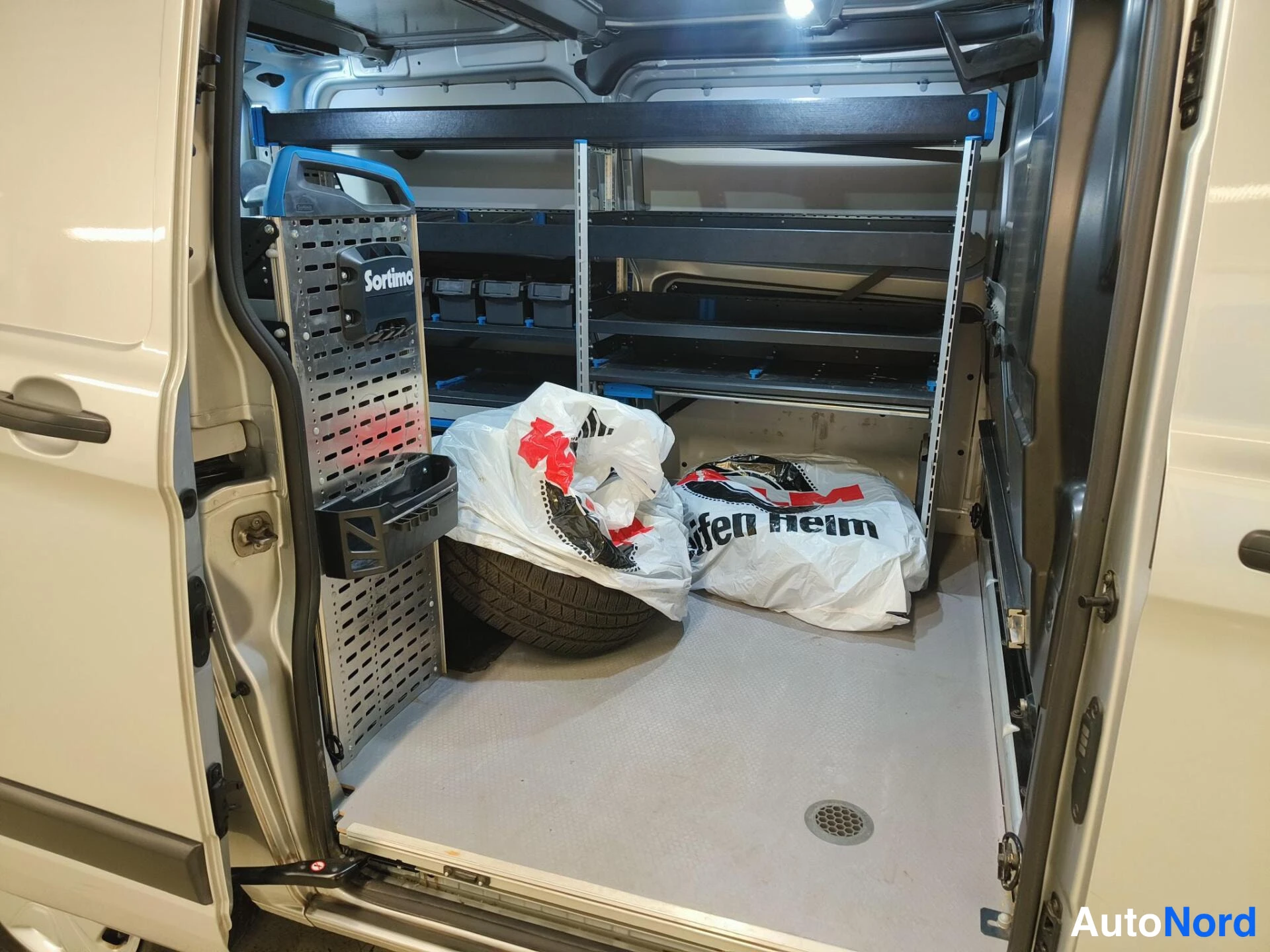 Muu: Pakettiauton kuormatila takaa päin kuvattuna: vasemmalla Sortimo-teline ja työkalusysteemi, lattialla varaosa ja valkoiset muovikassit, lattialämmitys- ja kaste
