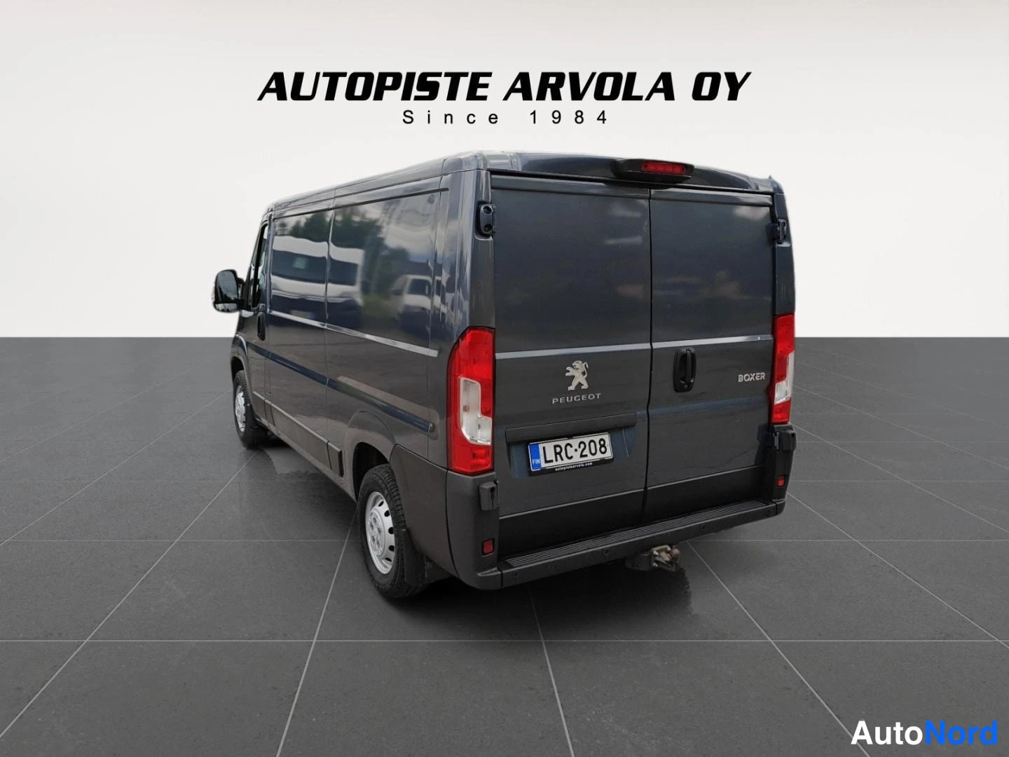 Takaviisto: Tummanharmaa Peugeot Boxer -pakettiauto takaviistolta, rekisterikilpi, auton takana teksti Autopiste Arvola Oy Since 1984.