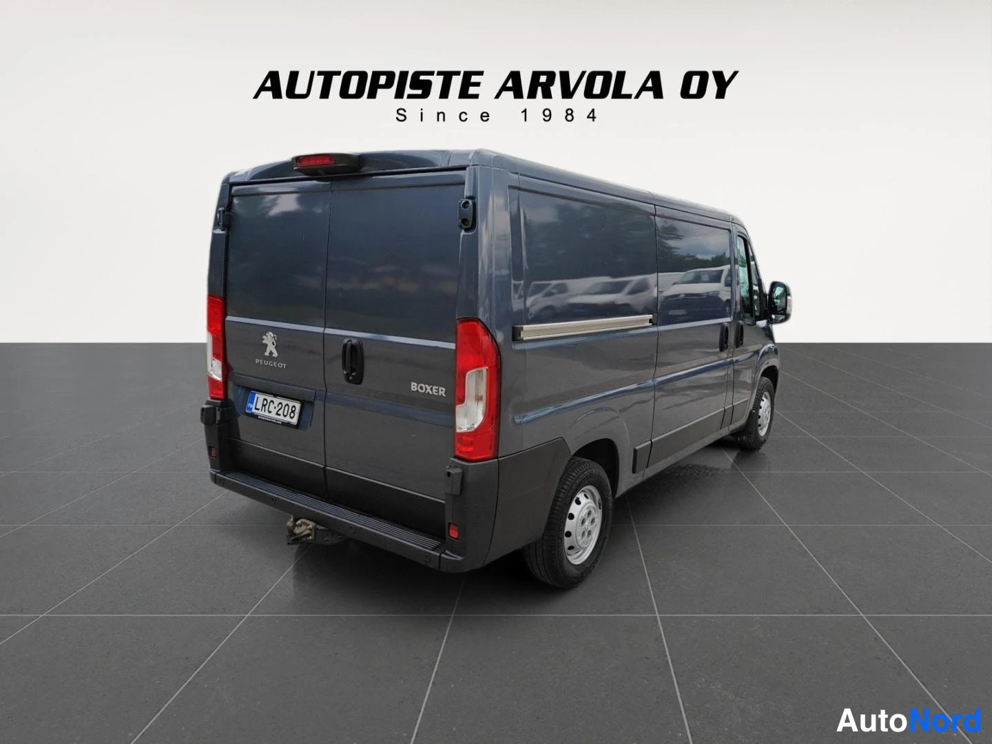 Takaviisto: Tummanharmaa Peugeot Boxer -pakettiauto takaviistolta toisesta kulmasta, rekisterikilpi, studiossa harmaata lattiaa vasten.
