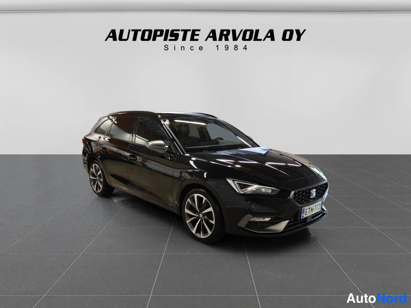 Etuviisto, nokka oikealle: Musta Seat Leon Sportstourer farmariauto valkoisella näytteistystyylillä kyljestään ja etupäästään kuvattuna. Auton etuvalot ovat kirkkaat ja etusuoja on
